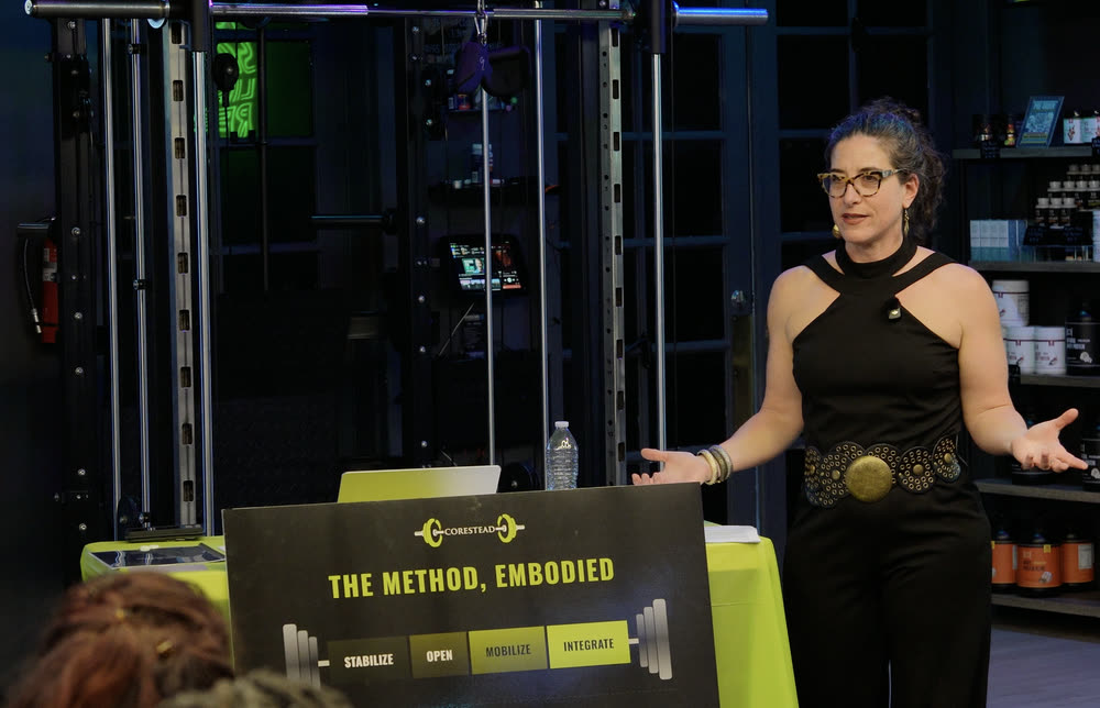 Emily Frydrych speaking to a seated audience at a Lifting From Within workshop, gesturing beside the Corestead lectern sign.